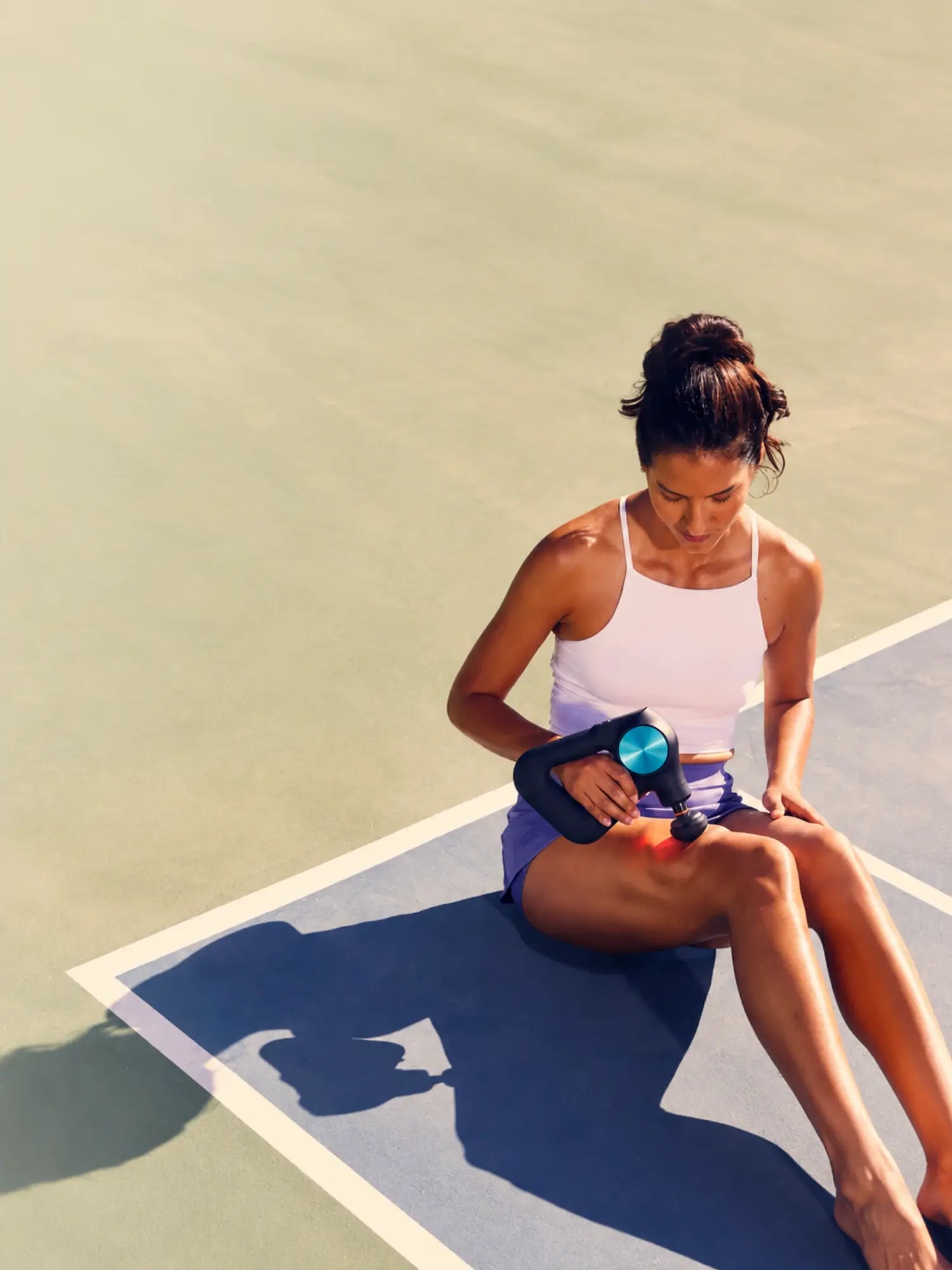 A woman sits on a tennis court holding a small handheld massage or heat device against her leg. She is wearing a white tank top and purple athletic shorts, with the court surface and her shadow visible in bright sunlight.