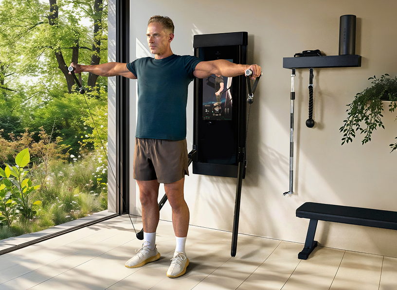 A man stands in a bright home gym using a wall-mounted cable crossover exercise machine, with a window showing green foliage outside and a bench and shelf with accessories nearby.