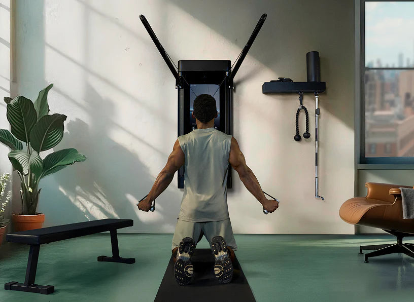 A man from behind using a wall-mounted pull-up/underbar row exercise machine in a modern, sunlit home gym with a bench, potted plant, and office chair.