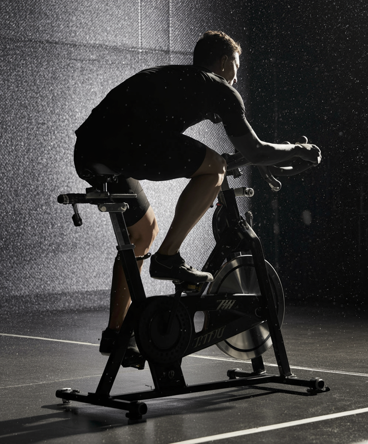 A person riding a black indoor cycling machine in a dark studio setting with dramatic lighting and visible water droplets in the air.