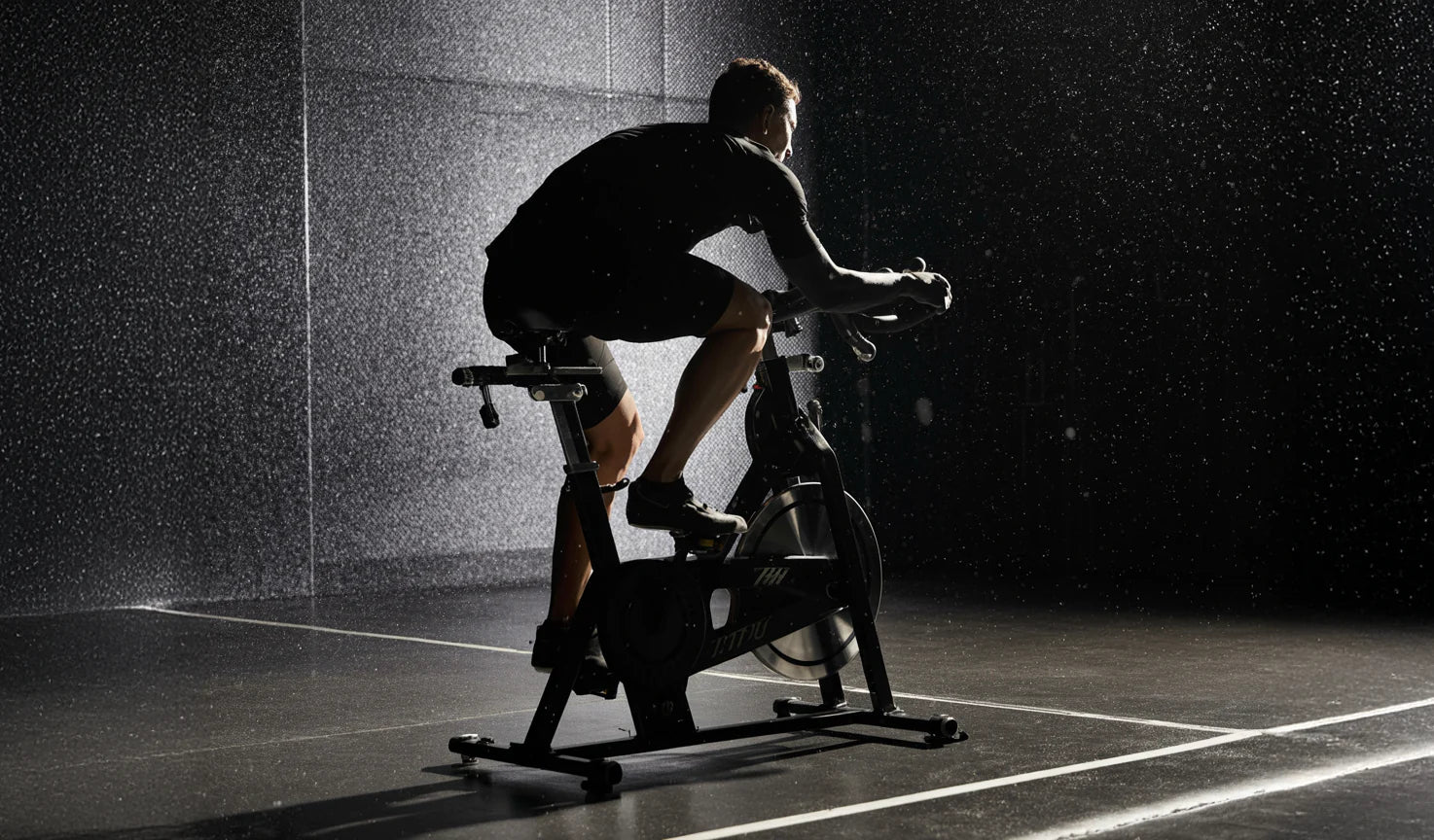 A person riding a stationary indoor bike in a dark, dramatic studio setting with water droplets in the air.